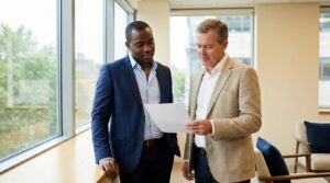 Financial planner and business owner standing at a bright office window reviewing a single printed page together.